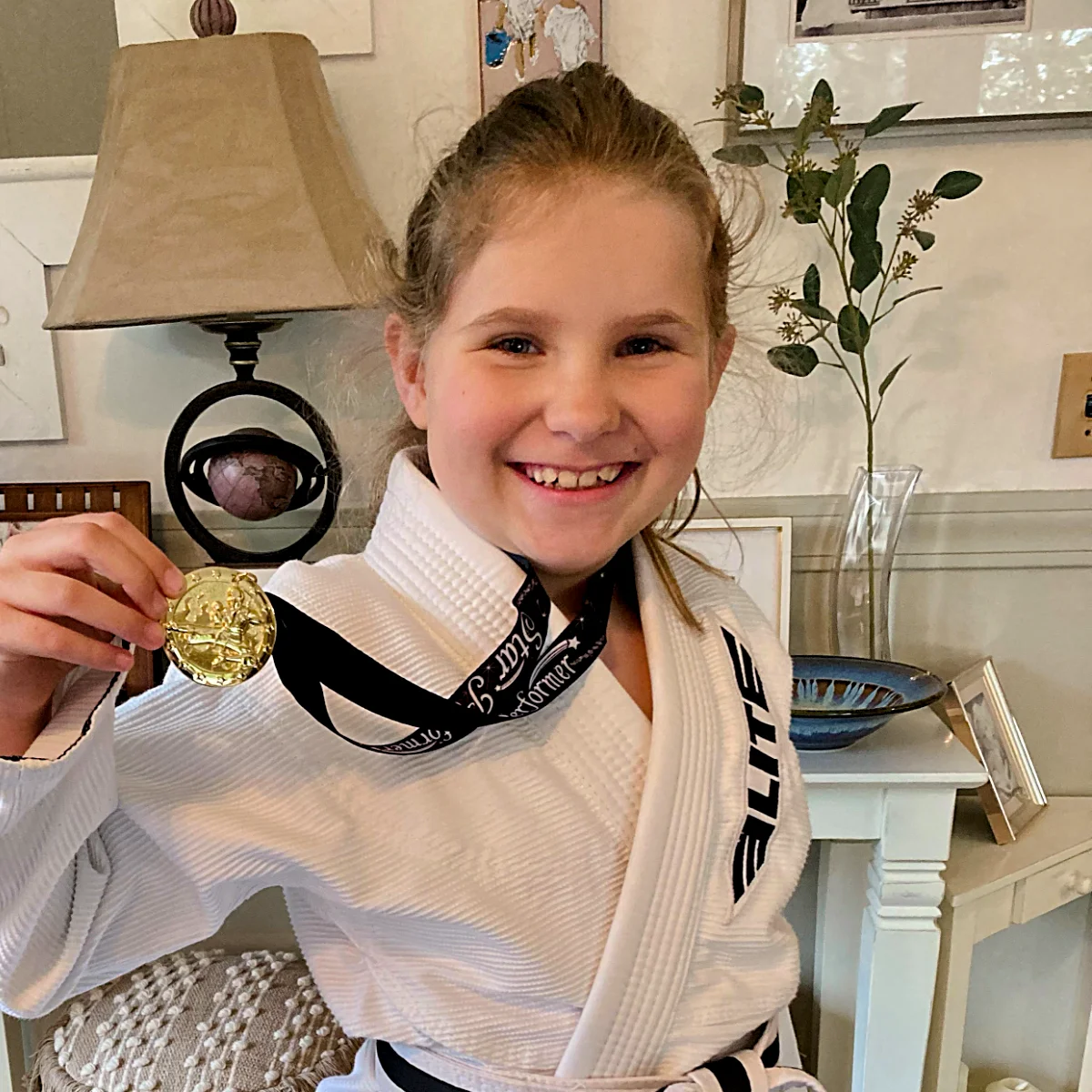 A smiling young girl in a white gi with a black belt proudly displays a gold medal awarded to her for achievement.