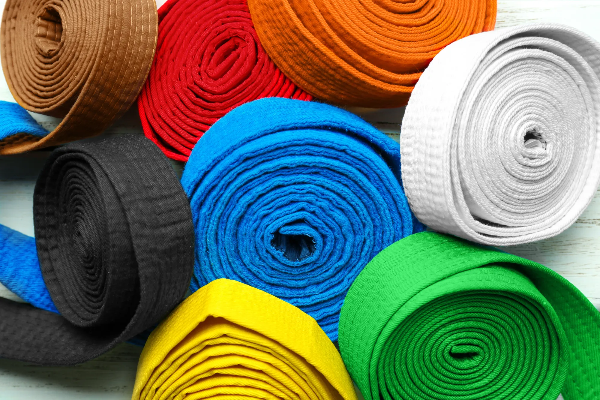 A close-up view of eight tightly rolled martial arts belts (obi) in various colors: white, black, red, blue, green, yellow, orange, and brown.