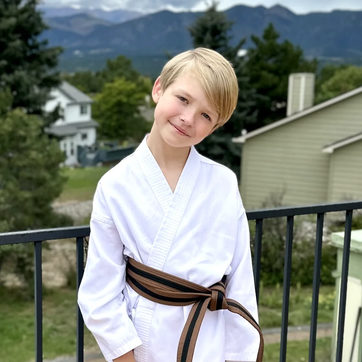 A young boy in a white karate gi and a brown belt smiles confidently while standing on a balcony. The background features houses, trees, and distant mountains.