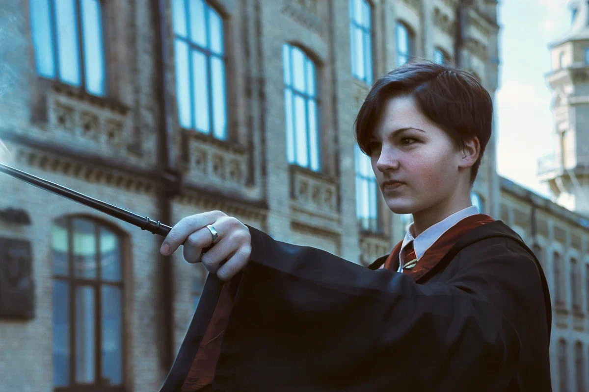 A student in a dark academic robe holds a wand emitting a bright blue magical burst. The focused person stands before an ornate brick university building.