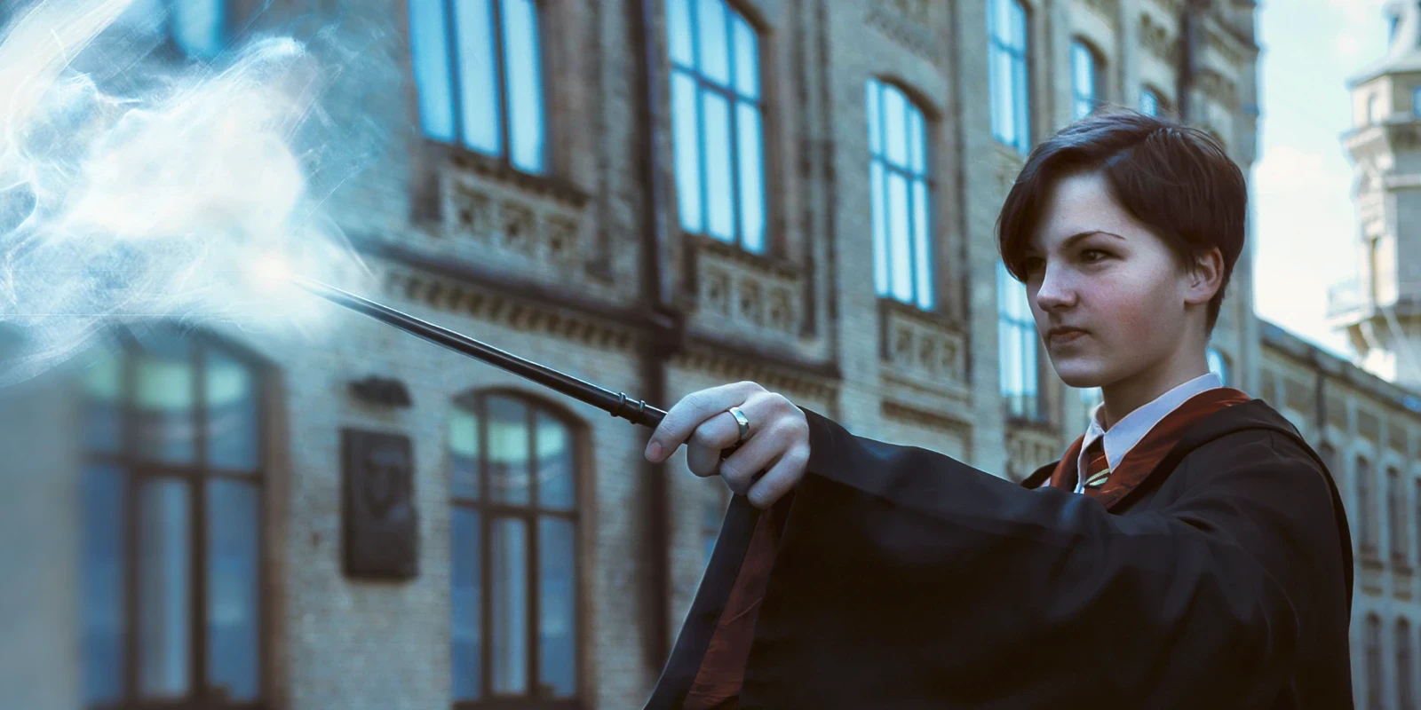 A student in a dark academic robe holds a wand emitting a bright blue magical burst. The focused person stands before an ornate brick university building.