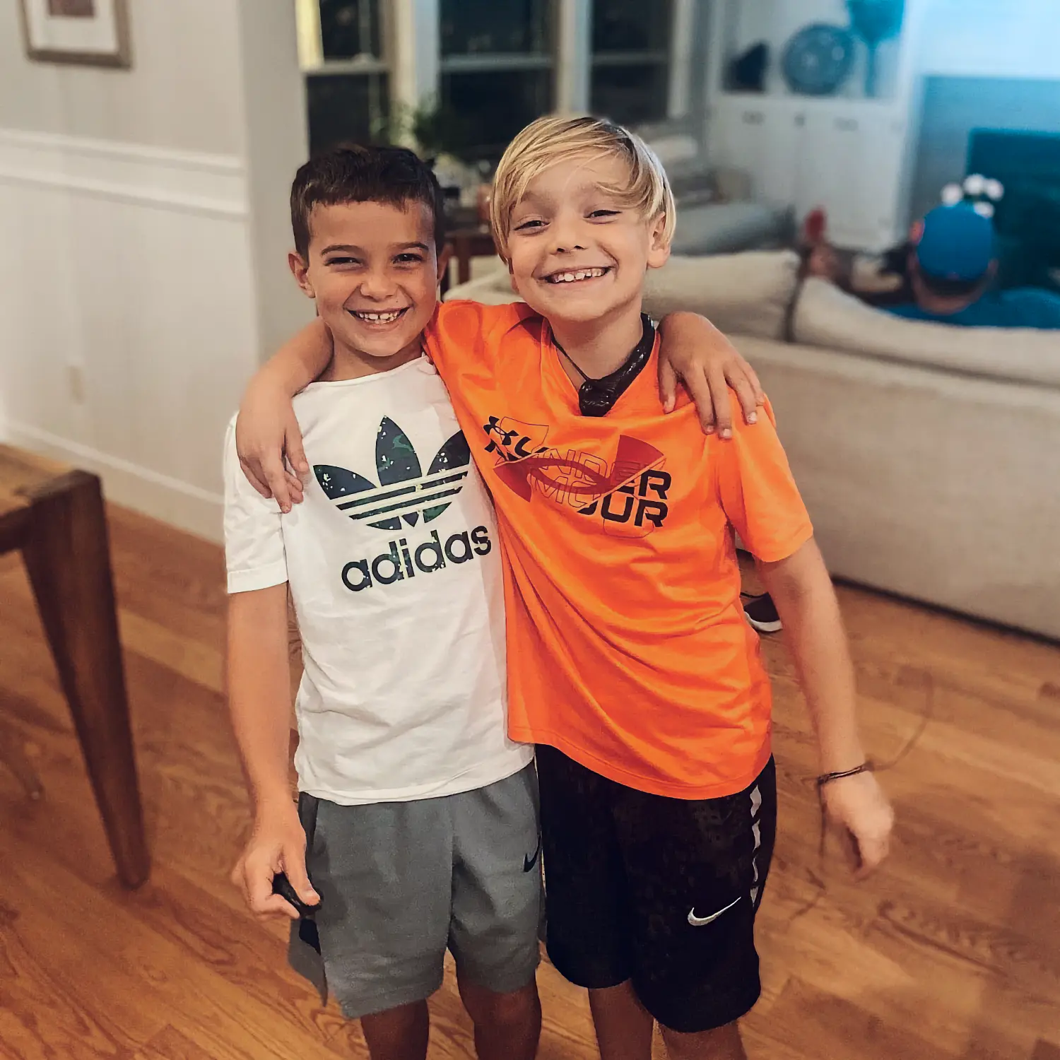 Two happy young boys in athletic clothes are embracing and smiling broadly at the camera inside a living room with wooden floors.
