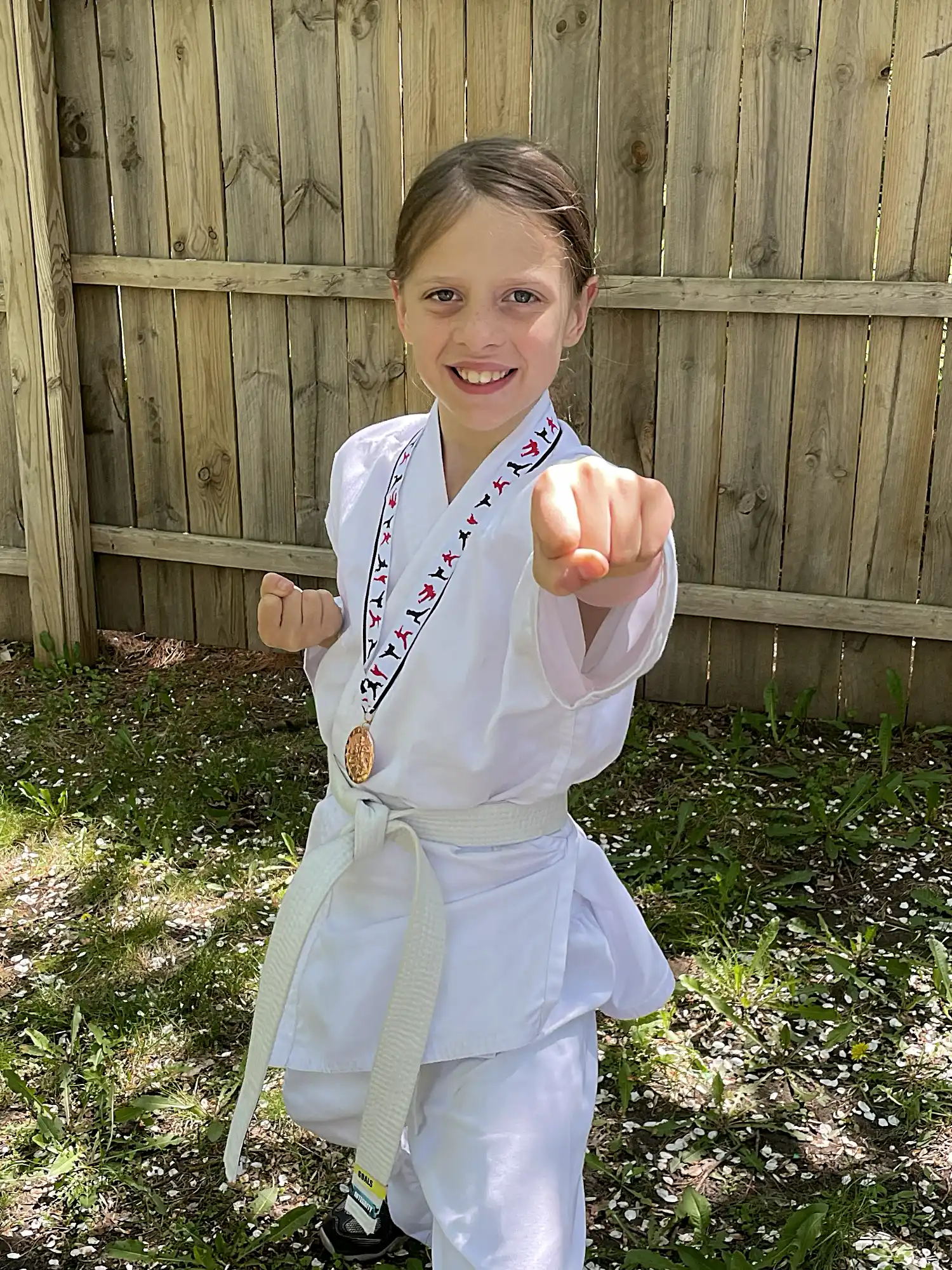A young girl in a white karate gi smiles confidently while executing a forward punch. She wears a medal around her neck, standing by a wooden fence.
