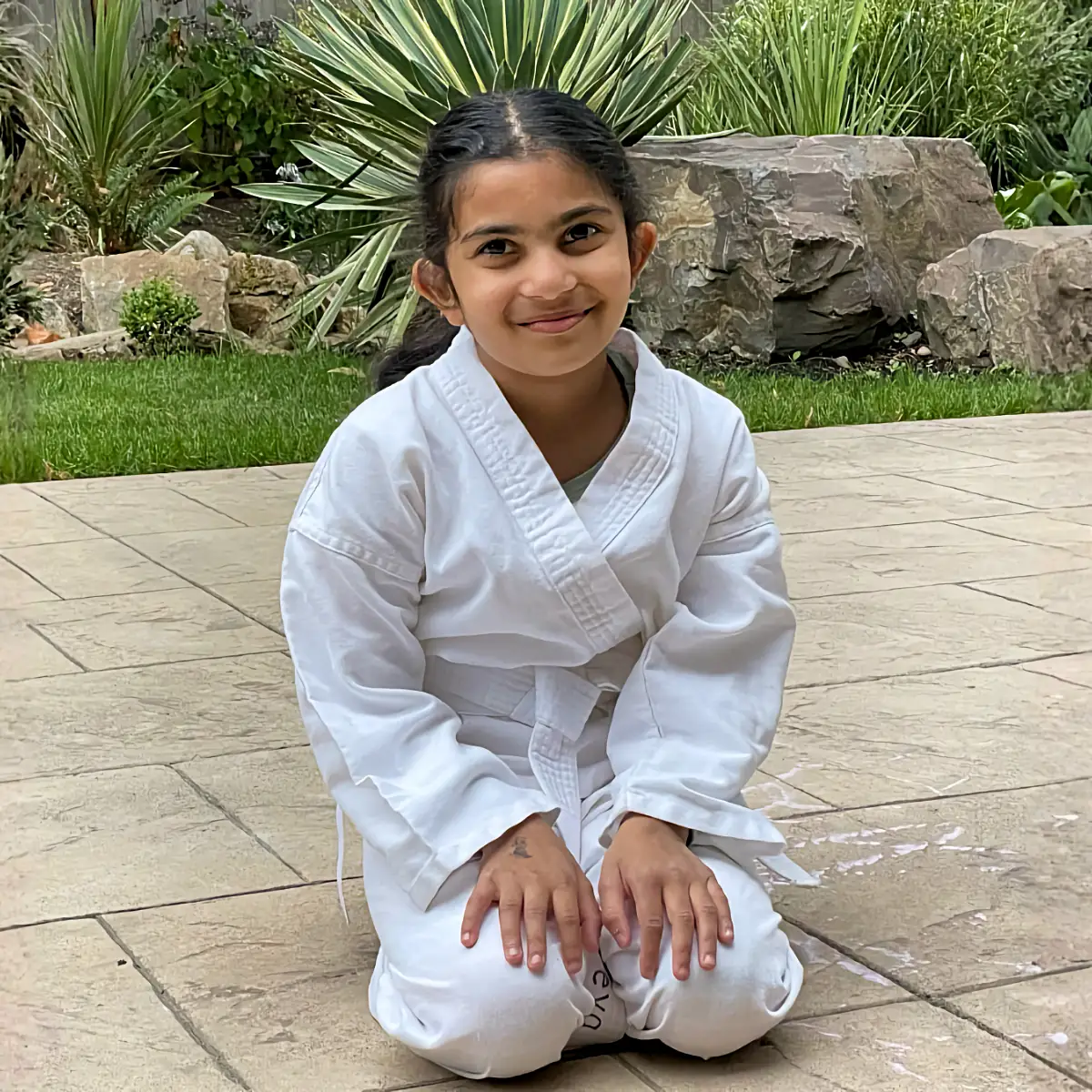 A young girl in a white karate gi kneels outside on a stone patio with a large smile. The background is a lush garden with rocks.