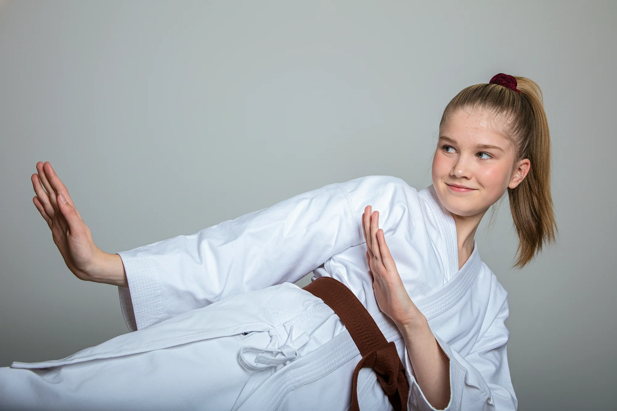 A young female karate student wearing a brown belt executes a side block, striking a dynamic pose. She is smiling confidently.