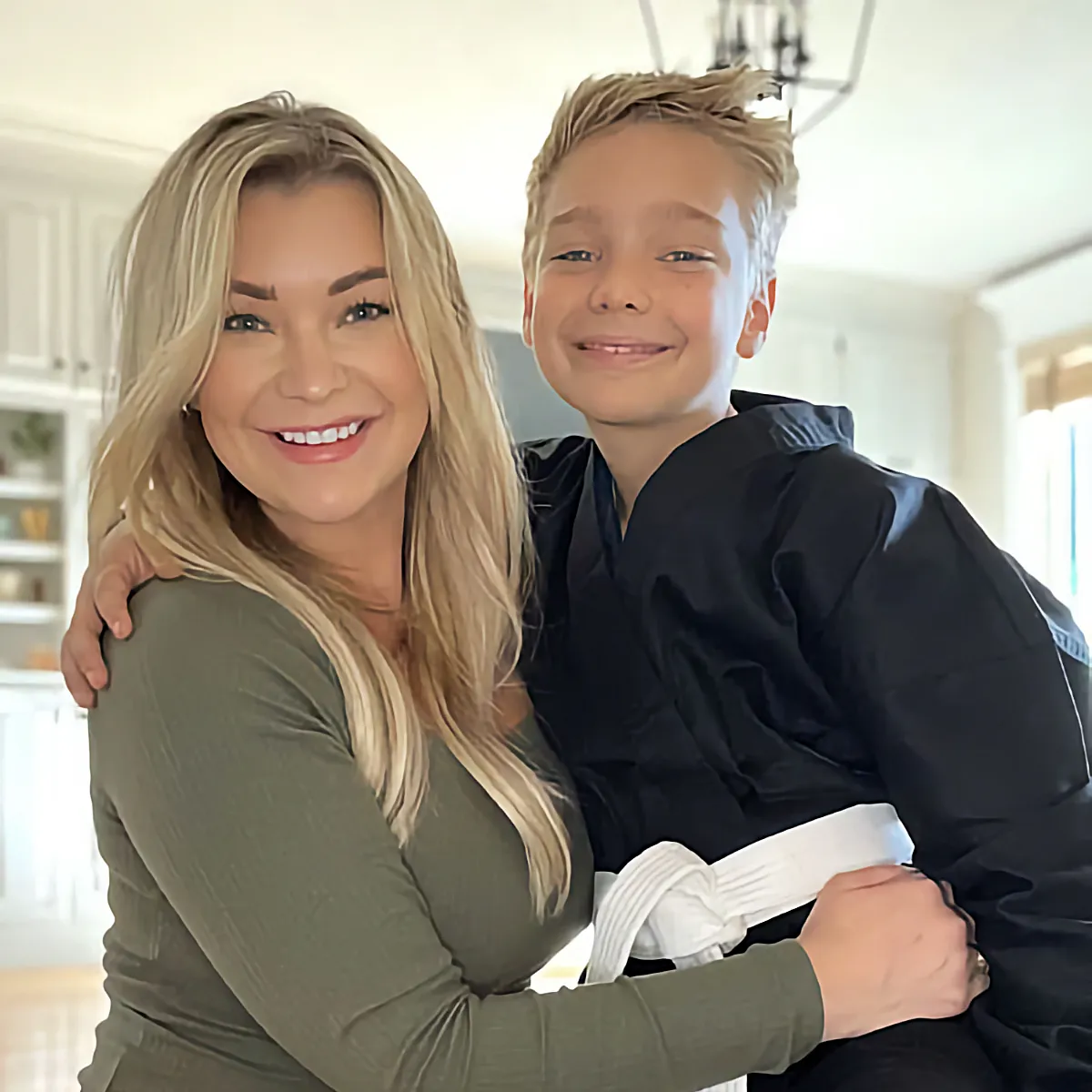 A smiling mother in a green shirt hugs her young son who is wearing a black karate uniform with a white belt.