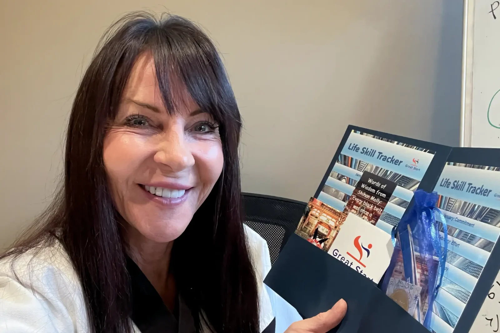A smiling Jan Stockton, a karate instructor, holds an open Great Start Life Skill Tracker folder containing program cards and a small blue pouch of materials.