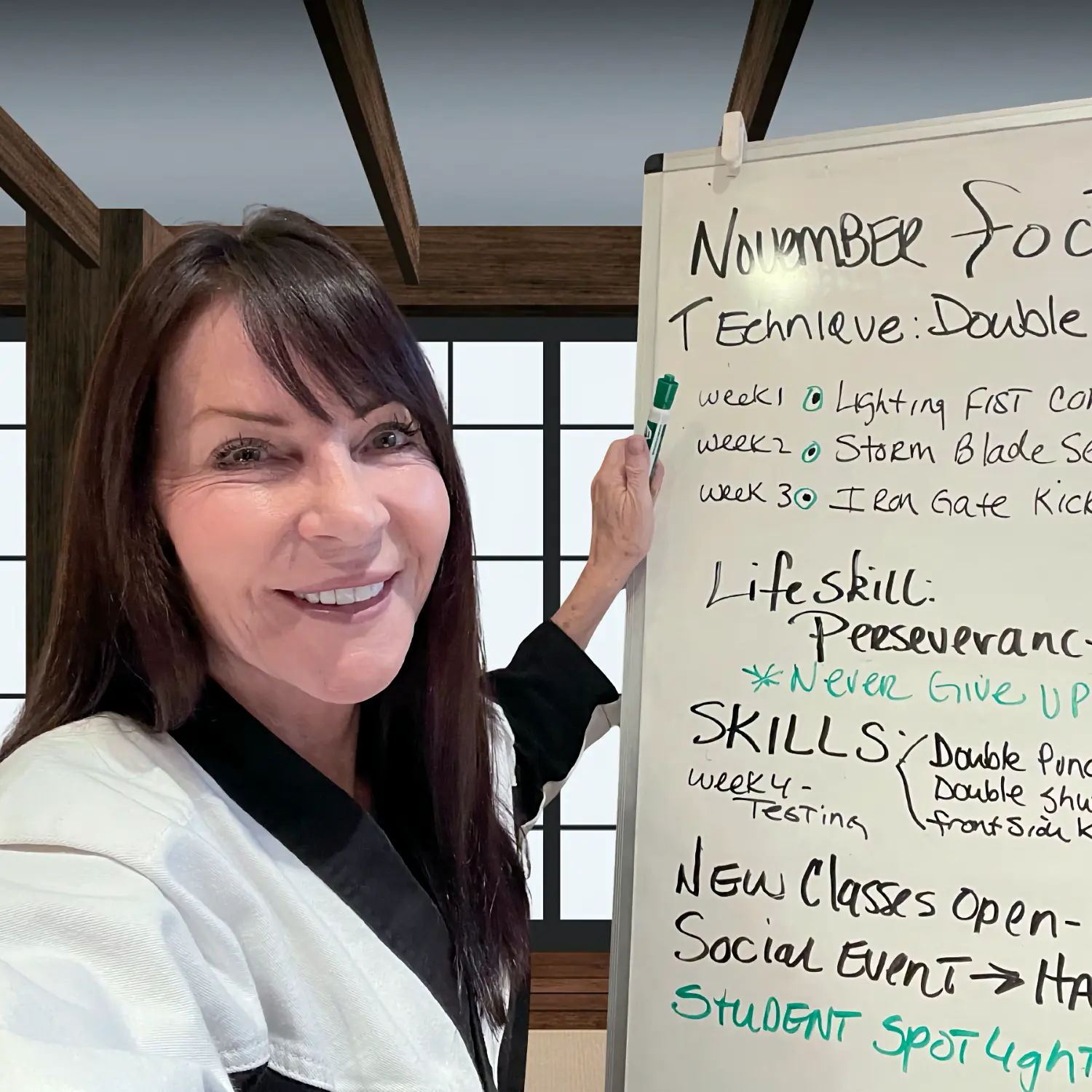 Karate instructor Jan Stockton smiles brightly next to a whiteboard listing the November focus, including techniques and the life skill, 'Perseverance.'
