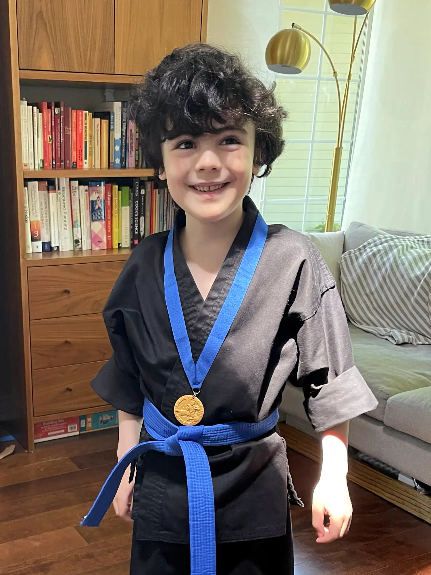 A young boy wearing a black karate uniform with a blue belt smiles proudly while displaying a gold medal in his living room.