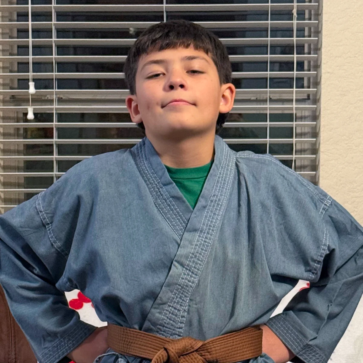 A confident boy in a blue martial arts gi and brown belt stands with his hands on his hips, looking proud and ready.