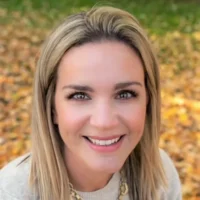 Andrea Mertins, a blonde woman, looks forward with a cheerful, open smile. She is wearing a light sweater outdoors against yellow autumn foliage.