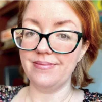 Audrey Sargent, with auburn hair and black and teal glasses, looks directly at the camera with a friendly, professional expression.