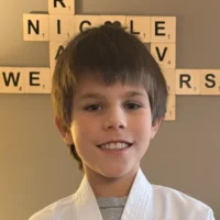 Carson, a young boy in a white karate uniform, smiles confidently against a gray wall decorated with large wooden Scrabble tiles.