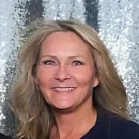 Laura Rude, a woman with blonde hair, smiles warmly while posing for a photo against a sparkling silver backdrop.