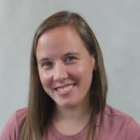 A headshot of Nicole P., a woman with brown hair wearing a pink shirt, looking directly at the camera with a cheerful, genuine smile.