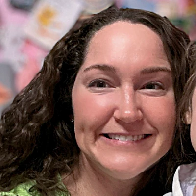 A woman with dark, curly hair smiles widely, showing a friendly and enthusiastic expression.