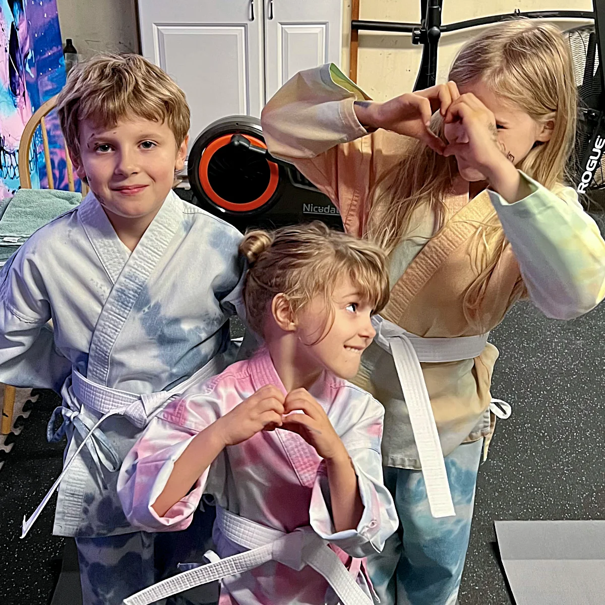 Three smiling kids in tie-dye karate uniforms show off their white belts. Two of the children are playfully forming heart shapes with their hands.