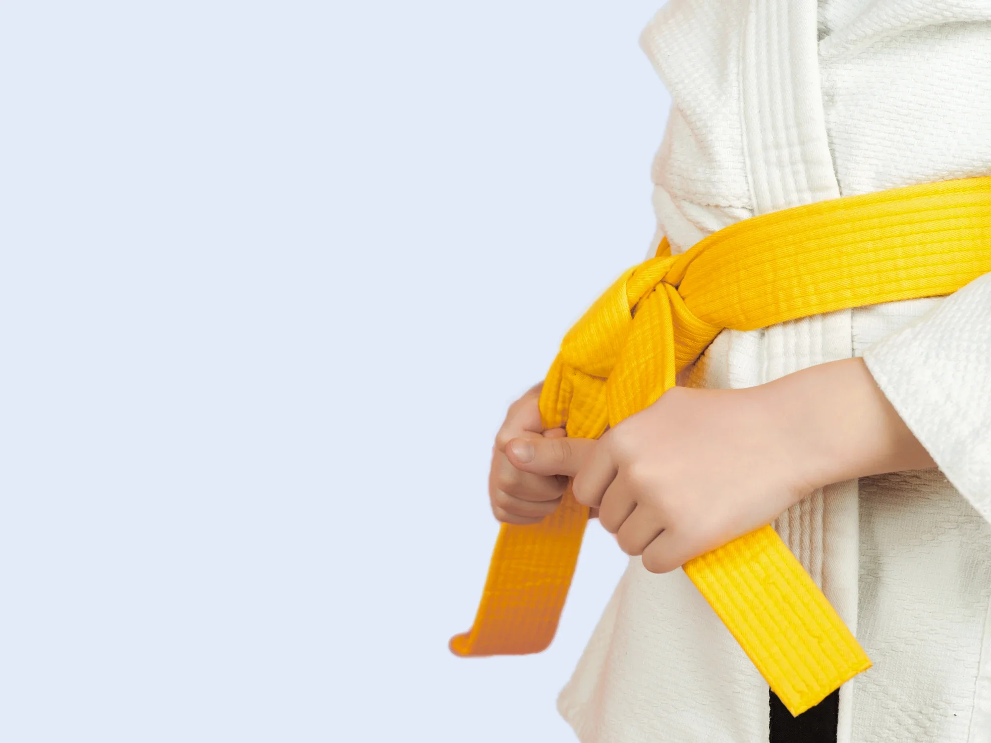 A kid in a white martial arts uniform adjusts their bright yellow belt with both hands against a light background.