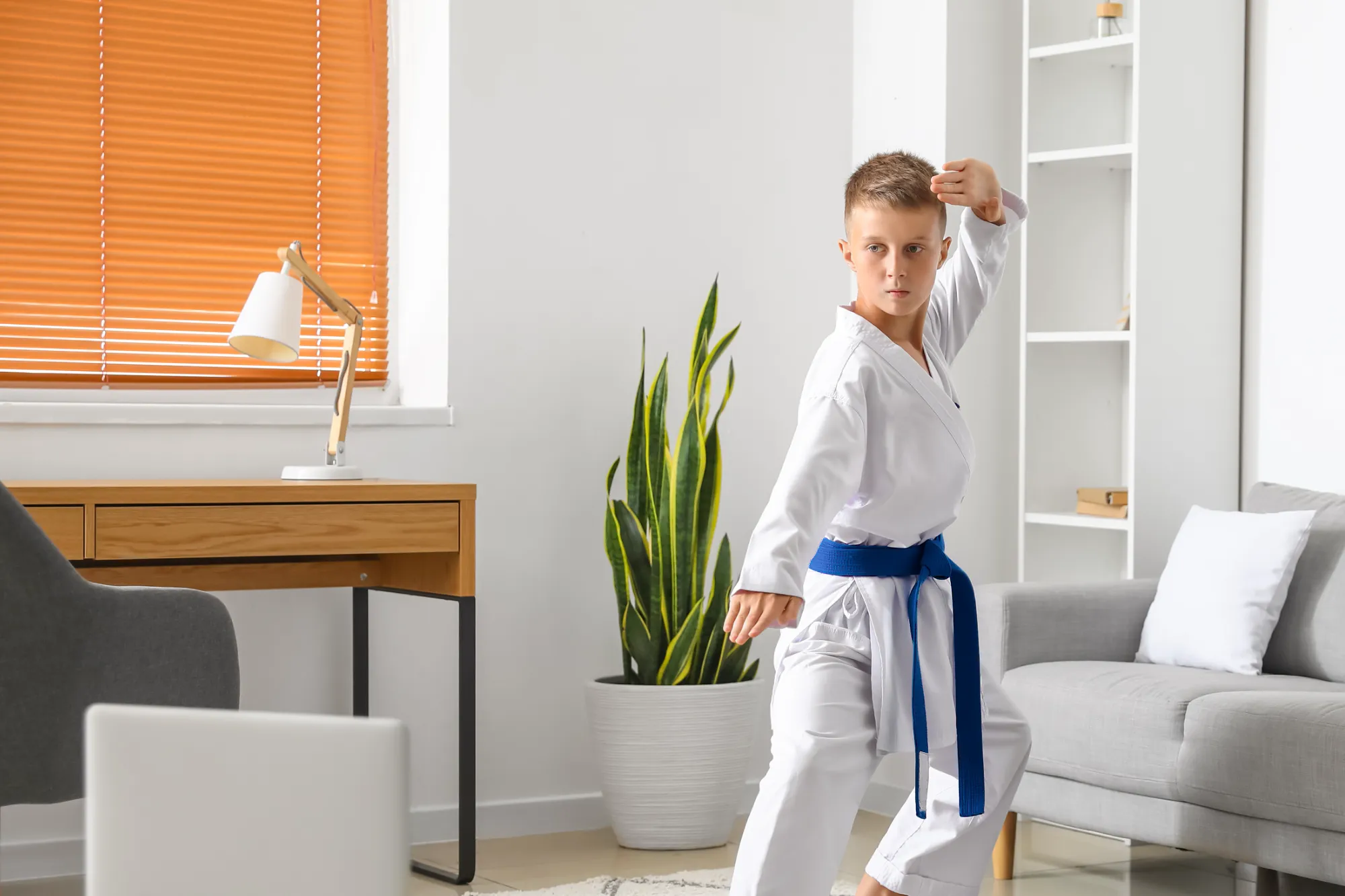 A focused boy wearing a white karate uniform and a blue belt performs a stance in a bright living room, practicing an online martial arts class via the laptop visible nearby.