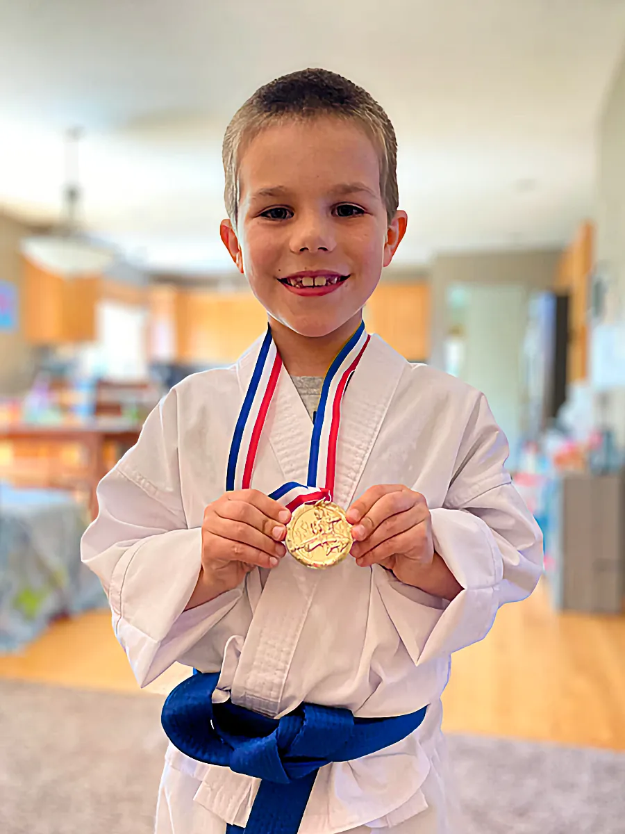 A happy kid in a white karate uniform and blue belt holds up a gold medal on a ribbon, smiling proudly.