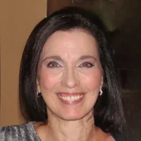 A portrait of Tina M., a woman with long dark hair, wearing earrings and a gray top, smiling warmly.