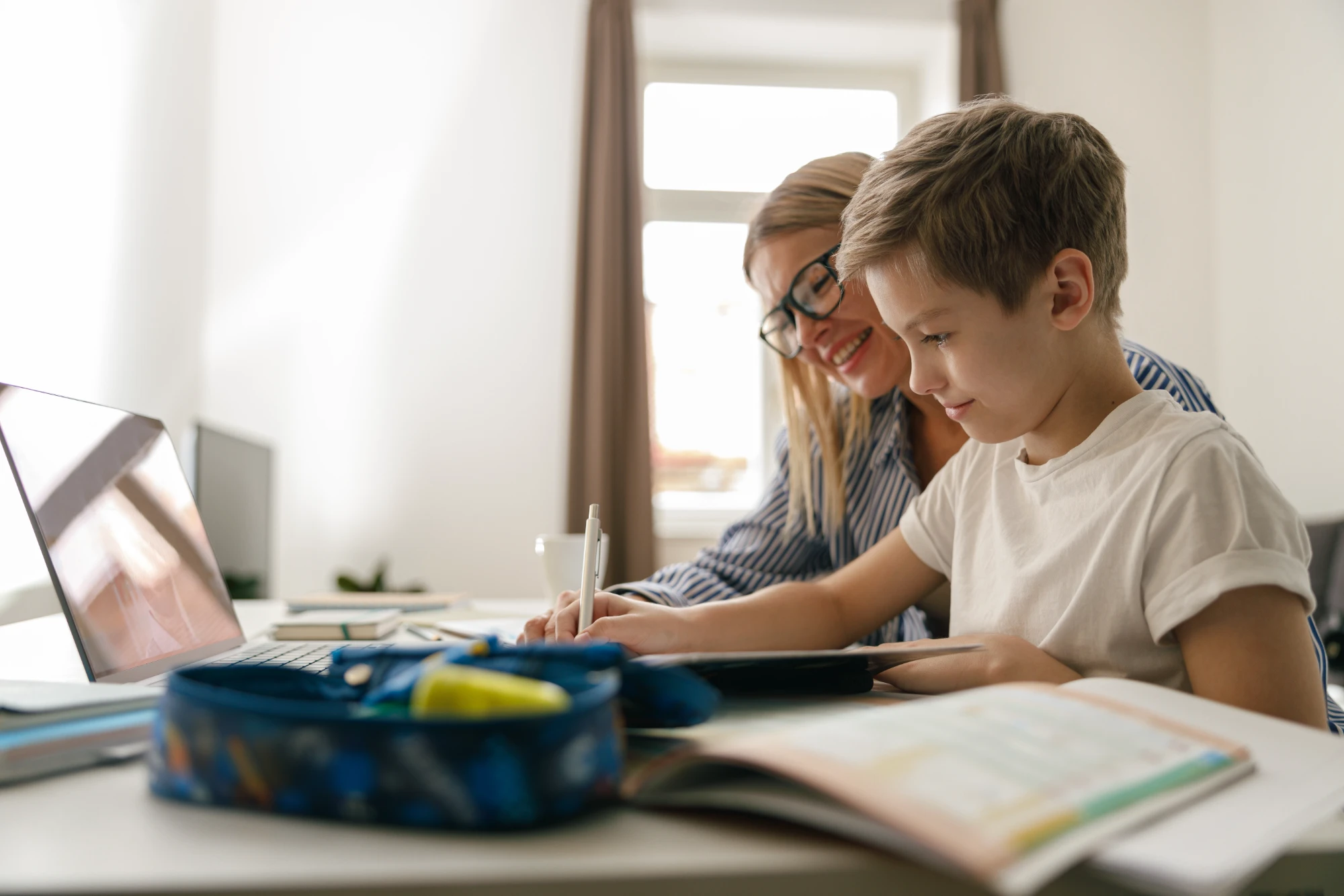 A smiling woman with blonde hair and glasses helps a young boy with light hair study at a bright desk, writing on a notebook next to an open laptop.
