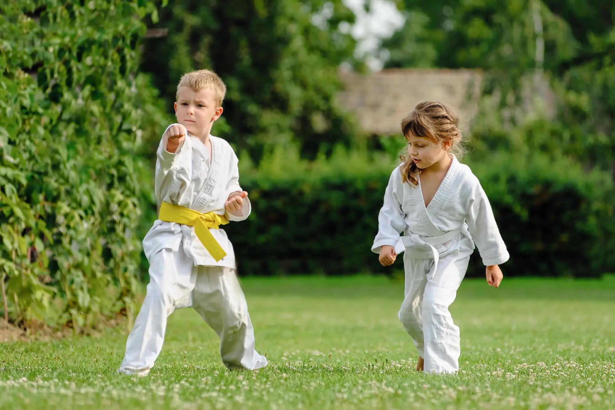 A boy with a yellow belt throws a focused punch while practicing karate outdoors on a lawn with a girl in a white gi.