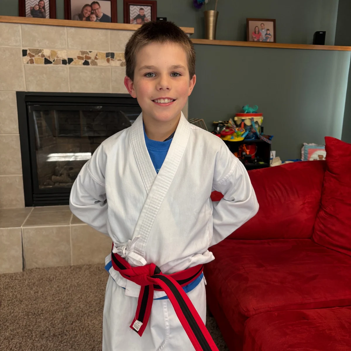 A young boy in a white martial arts uniform with a red and black belt stands in a proud stance and smiles in his living room.