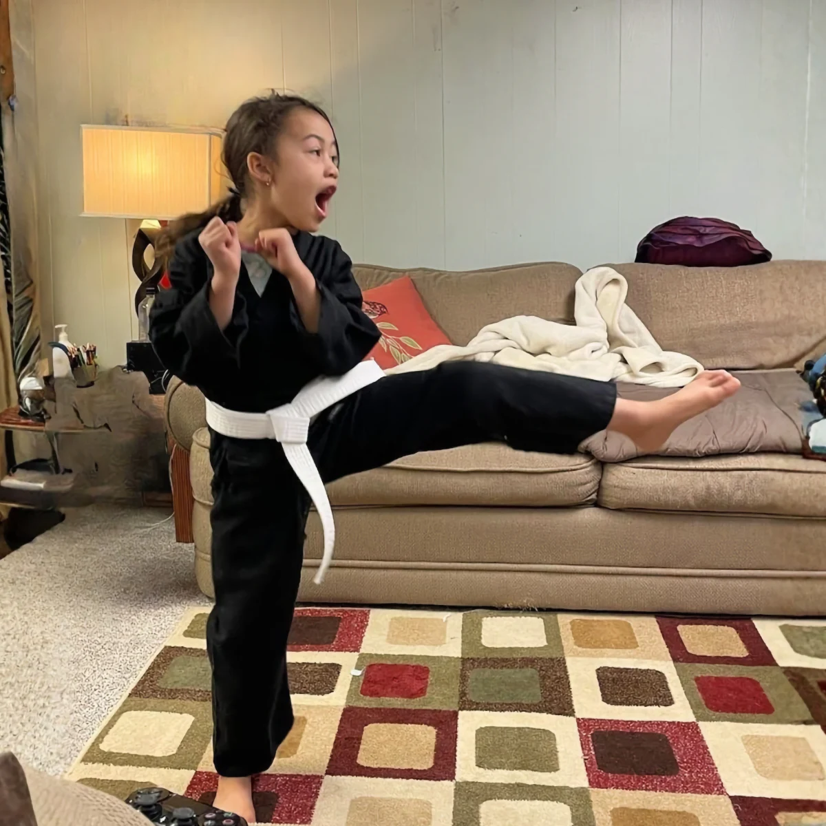 A young girl wearing a black karate uniform and white belt executes a high kick in a living room, demonstrating focus and power.