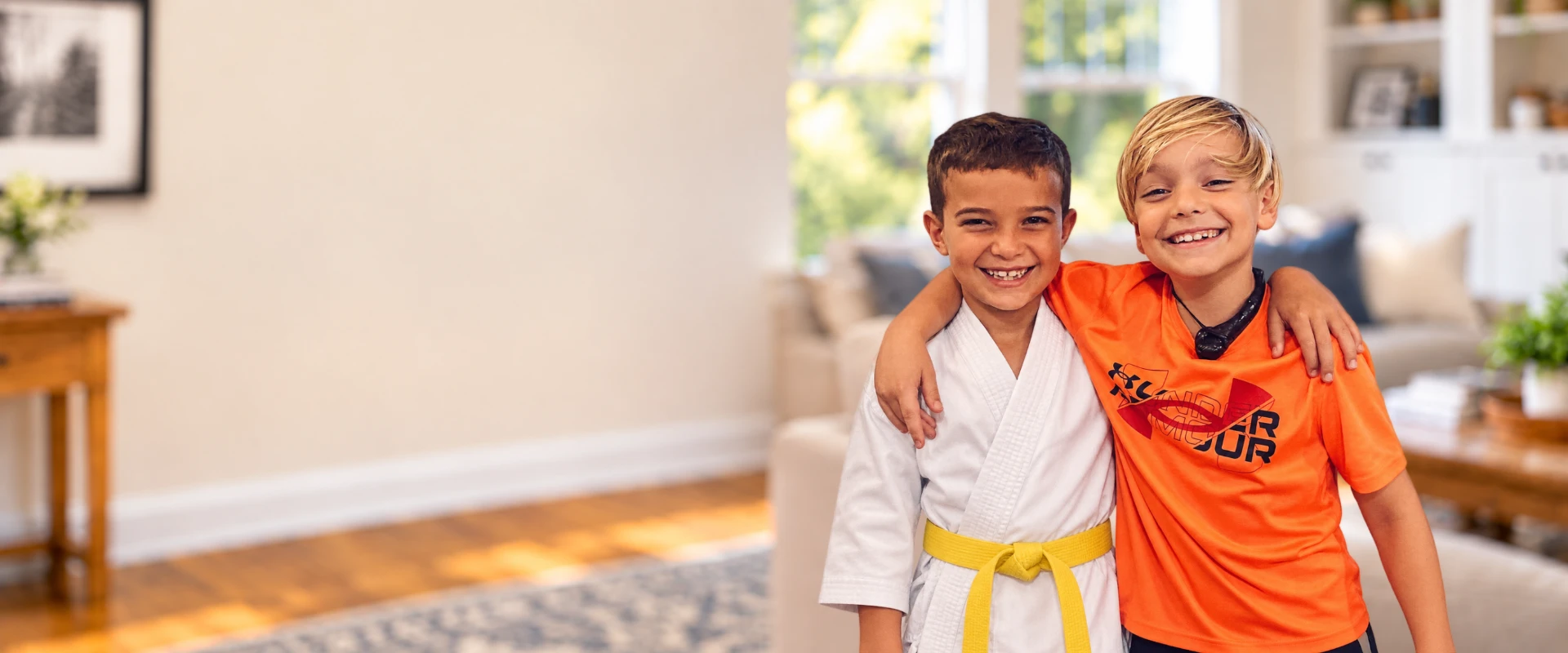 Two smiling boys stand shoulder-to-shoulder in a bright living room. One wears a white karate gi and yellow belt, and the other wears an orange athletic shirt.
