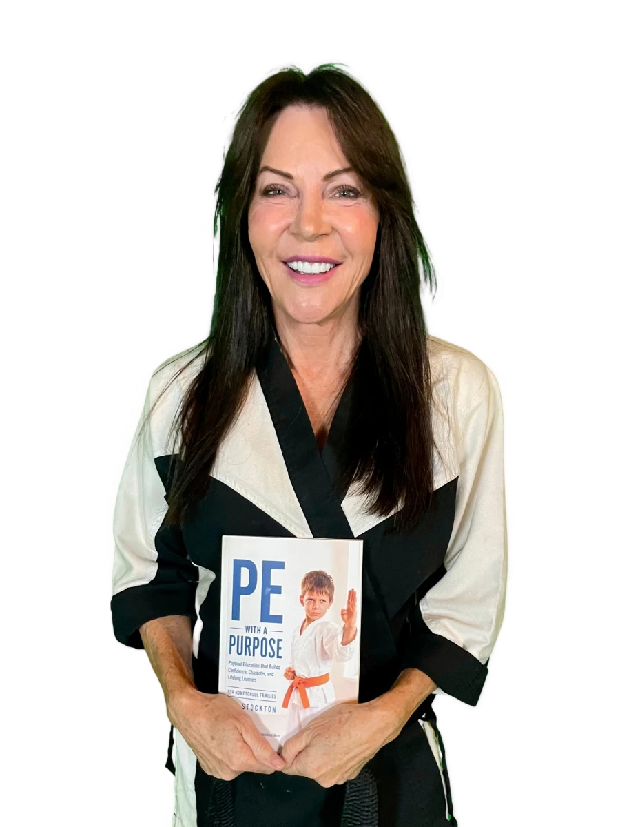 Martial arts instructor Jan Stockton smiles brightly, wearing a white and black uniform and holding her book, "PE with a Purpose."